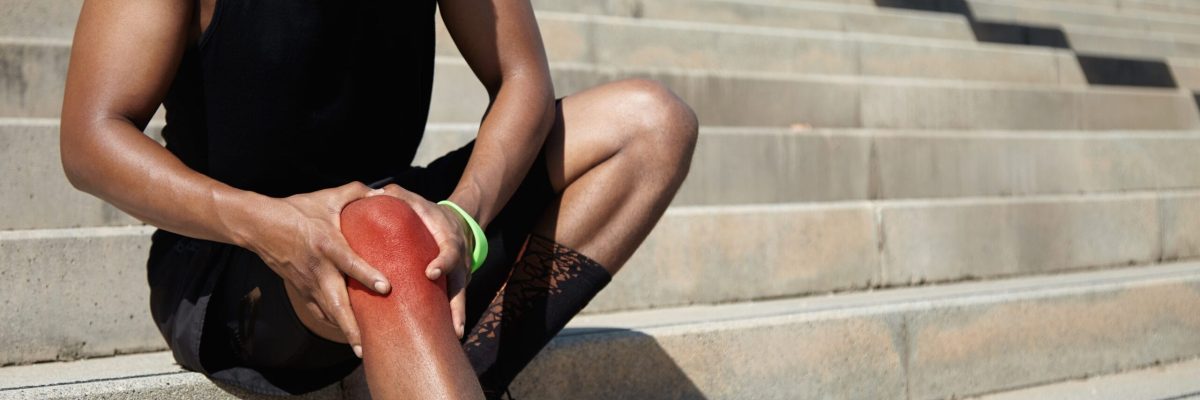 Cropped portrait of dark-skinned jogger in training socks and running shoes having sprain, clutching his injured knee, massaging it with both hands after intense workout in urban surroundings