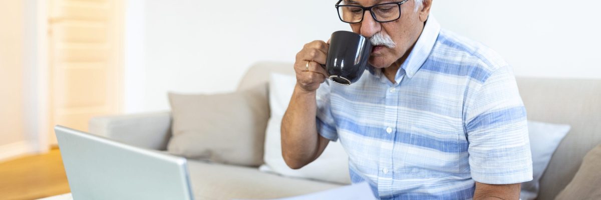 Focused grey-haired elderly man sit on couch reading bank notifications calculating domestic expenditures, concentrated modern mature male consider financial paperwork, pay bills on laptop online