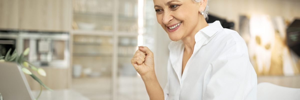 Successful mature female writer sitting at desk with portable computer, papers and cup of coffee, having happy facial expression because she managed to finish work in time, sending article to editor